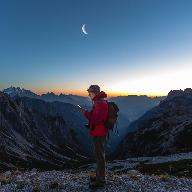 Hiker checking his mobile phone while enjoying the sunset and moonrise.