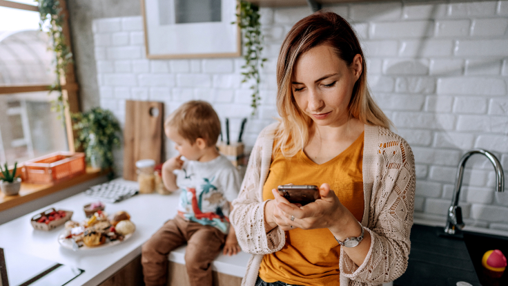 Woman checks her phone for a message while preparing a meal for her child.