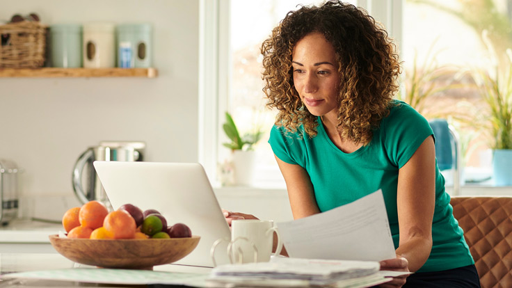 Woman looks at a laptop screen while shuffling papers.