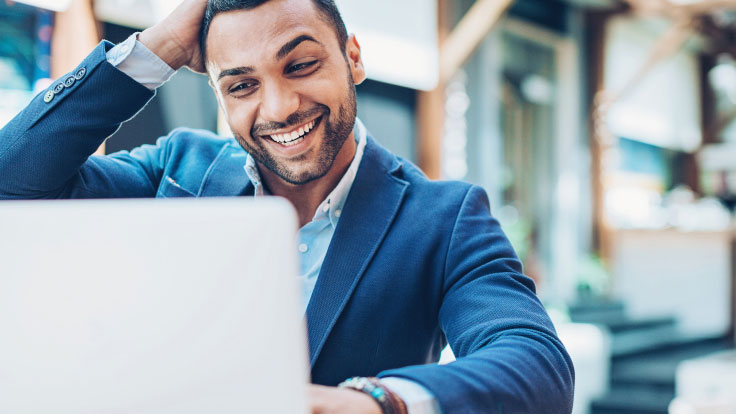 A smiling man sits in front of his laptop.