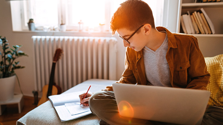 A teenage boy sits in his bedroom with a laptop while writing in a notebook.